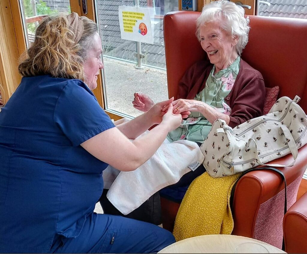 A nurse in blue scrubs holds the hands of an elderly woman seated in a chair, both smiling. The woman has a bag next to her and a blanket on her lap.