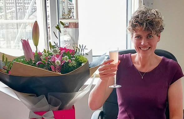 A Care Manager sits at a desk, smiling as she holds a glass of pink drink, with a large bouquet of flowers in a gift bag next to her.