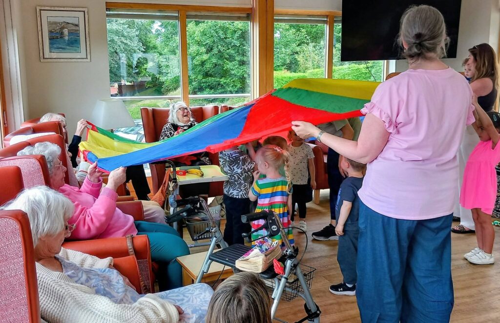 Children and elderly people play together in a brightly lit room, holding up a colourful parachute. Some elderly are seated whilst the children and adults stand, all joining in the activity.