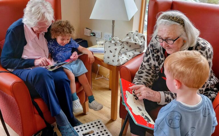 Two elderly women seated in armchairs read storybooks to a young girl and boy in a cosy room with a window, a table lamp, and a patterned bag.