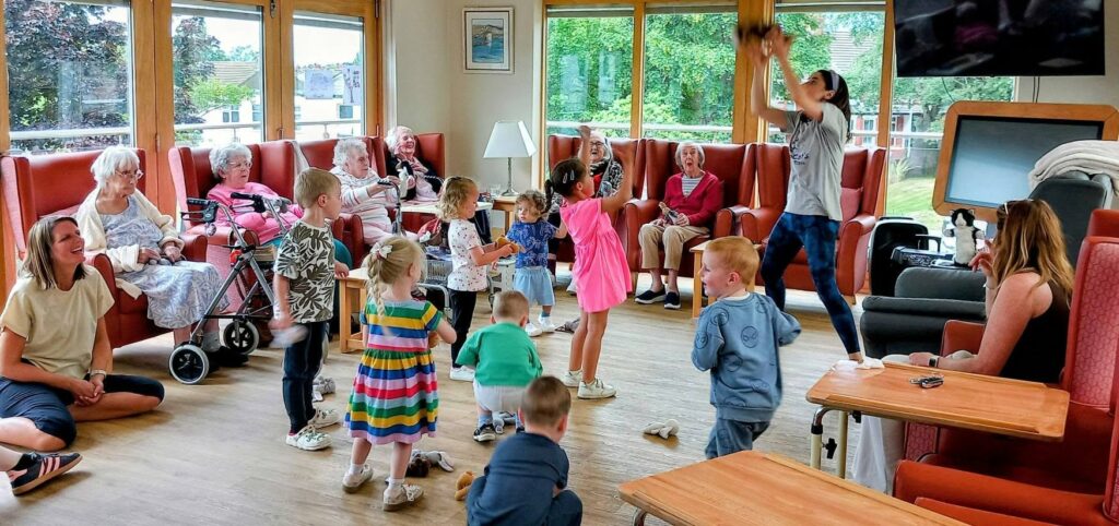 Children and elderly people interact in a bright communal room. Some children play on the floor as adults sit and observe, with one adult standing and raising their arms, perhaps leading an activity. Large windows reveal greenery outside.