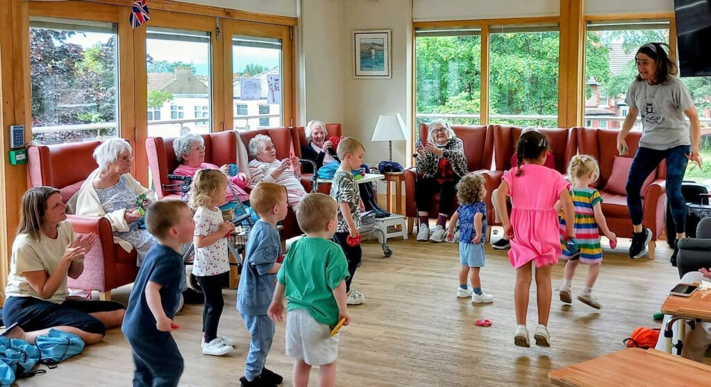 A group of young children dance and play in a bright room as several older adults watch from chairs, some smiling and clapping. An adult leads the activity. Large windows reveal greenery outside.