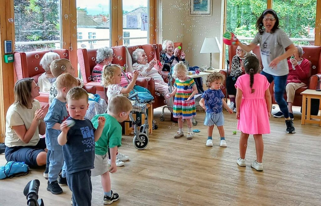 A group of young children and older adults sit and stand in a bright room, interacting and playing with bubbles whilst one adult supervises the activity.