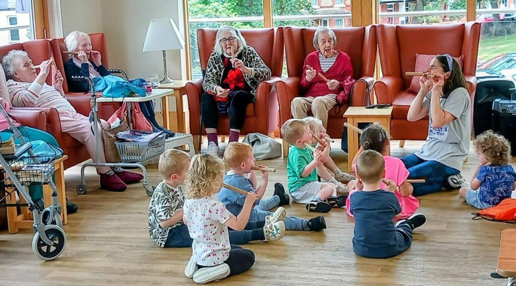 A group of children sit on the floor eating snacks in front of elderly women in armchairs in a bright room, indicating an intergenerational gathering.