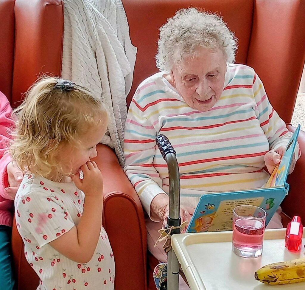 An elderly woman seated in an armchair reads a children’s book to a young girl standing next to her. In the foreground are a zimmer frame, a tray with a glass of pink drink, and a banana.