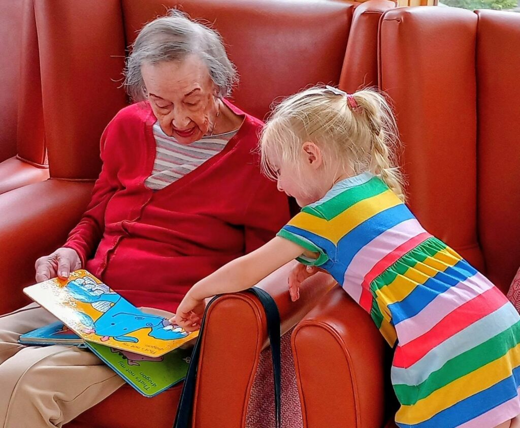 An elderly woman and a young girl sit together on an orange sofa, reading a colourful children's book. The girl points at something in the book whilst the woman holds it open.