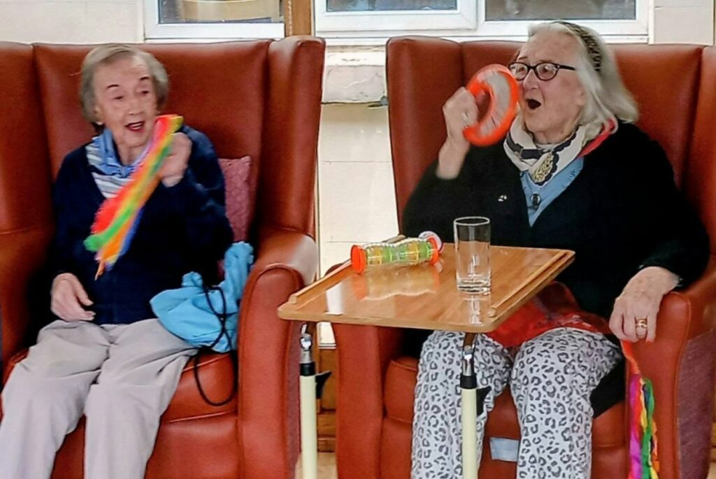 Two elderly women sit in armchairs, smiling and holding musical instruments. One waves a colourful scarf while the other plays a tambourine. A tray with a glass and a toy is placed in front of them.