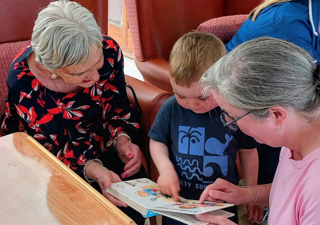 An elderly woman, a young boy, and an adult woman sit together indoors on red upholstered chairs, looking at a colourful book. The boy points at the pages as the adults watch attentively.
