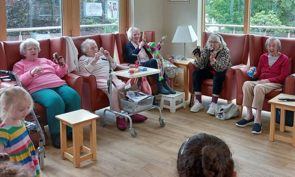 Five elderly women sit in a lounge holding soft toys, joined by two young children. The room has large windows, armchairs, small tables, and is filled with natural light.