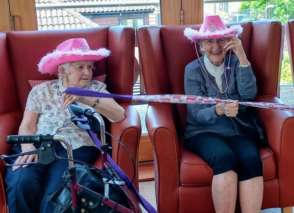 Two older women in pink cowboy hats sit on red chairs. One has a walking frame; the other smiles and holds a ribbon. They seem to be at an indoor event with large windows visible behind them.