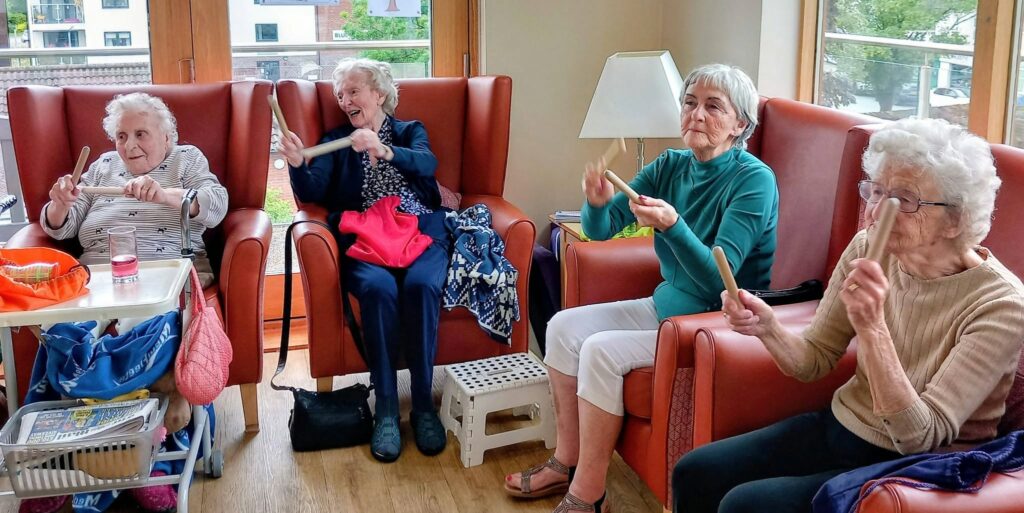 Four elderly women sit in armchairs in a well-lit room, each holding and playing wooden percussion instruments, looking engaged in a group activity.