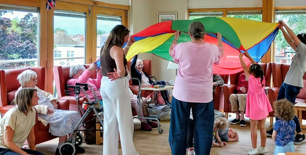 A group of adults and children in a brightly lit room hold a colourful parachute, while some elderly people are seated around them, watching.