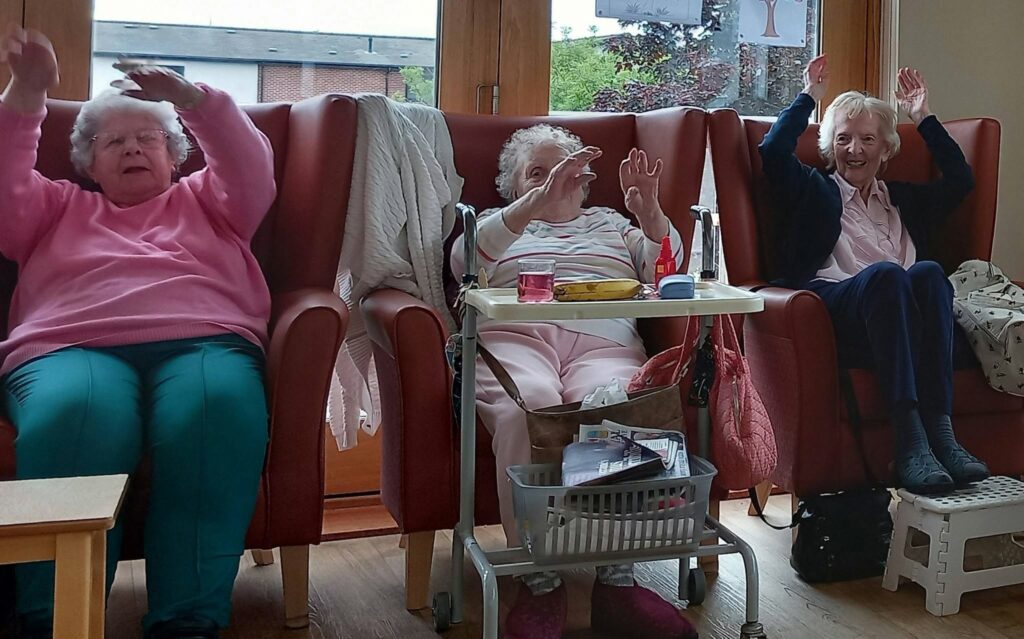 Three elderly women sit in armchairs with their arms raised, seemingly involved in a group activity. A tray with personal items rests before the woman in the centre. They are indoors near a window.