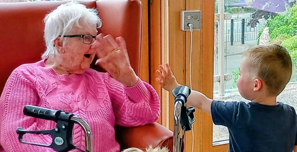 An elderly woman in a pink jumper and glasses sits in an armchair, raising her hand to greet a young boy standing nearby who is also lifting his hand. A walking frame is visible in front of her.