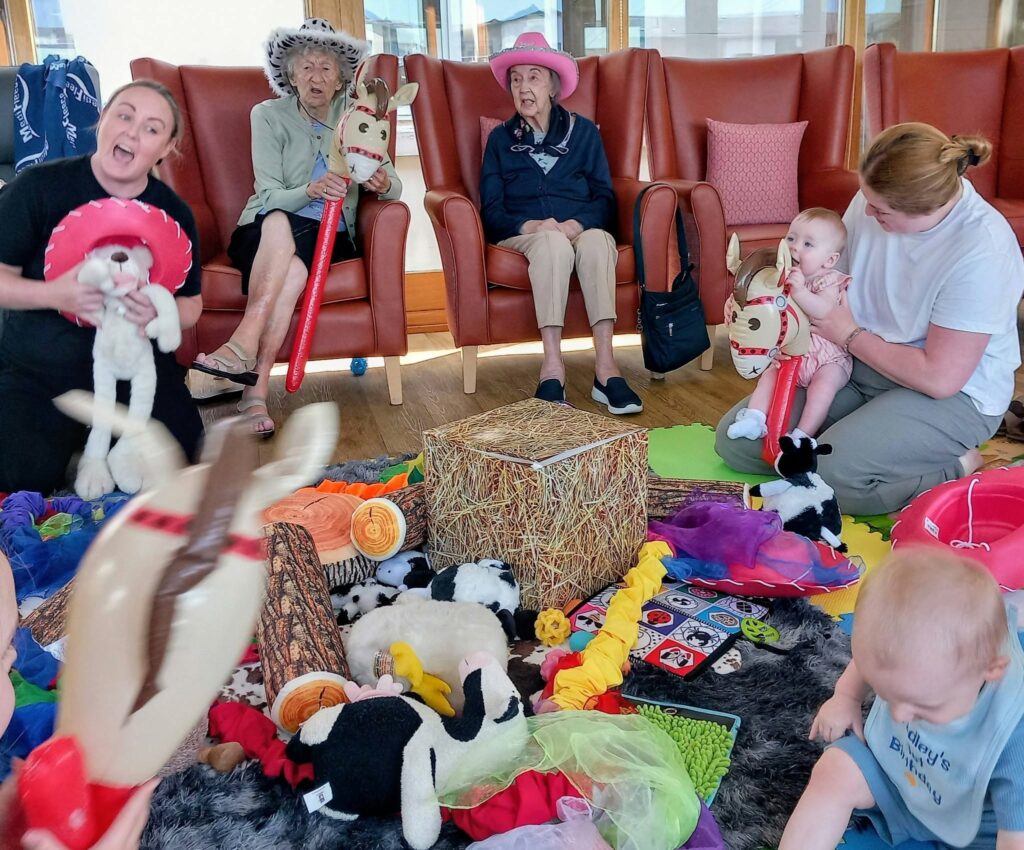 Elderly women, babies, and carers take part in a group activity with toy horses, soft toys, and colourful mats in a room furnished with armchairs.