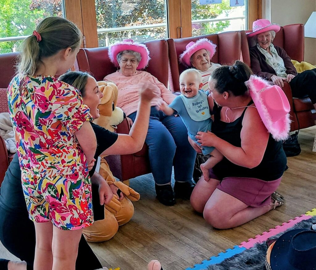 A group of older adults and children, some sporting pink cowboy hats, sit and kneel in a lounge, smiling and engaging with each other.