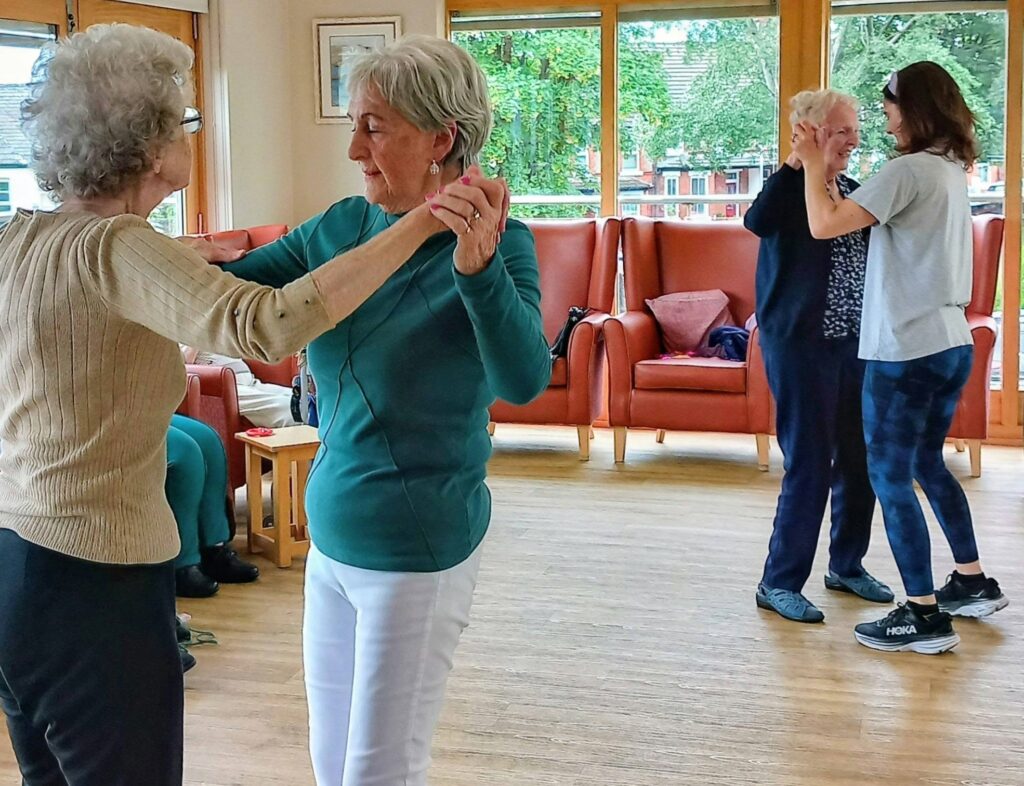 Four women, in pairs, are ballroom dancing in a bright room with large windows, wooden floors, and comfy orange chairs. They seem to be enjoying a social or exercise activity together.