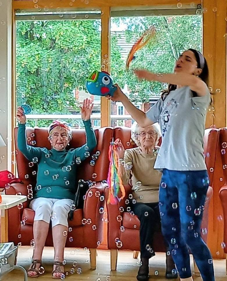 A woman blows bubbles with a toy as two elderly women sit in armchairs, one with her arms raised. The scene is set in a bright room with large windows and greenery visible outside.