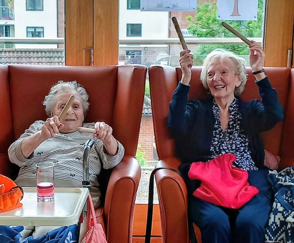 Two elderly women sit in armchairs, smiling and lifting wooden rhythm sticks above their heads, seemingly taking part in a musical or leisure activity indoors.