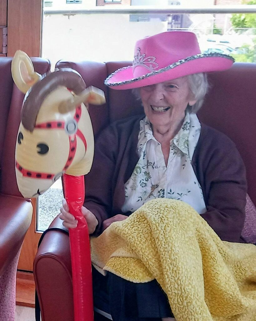 An elderly woman in a pink cowboy hat sits in an armchair, smiling whilst holding an inflatable toy horse. A yellow blanket rests on her lap.