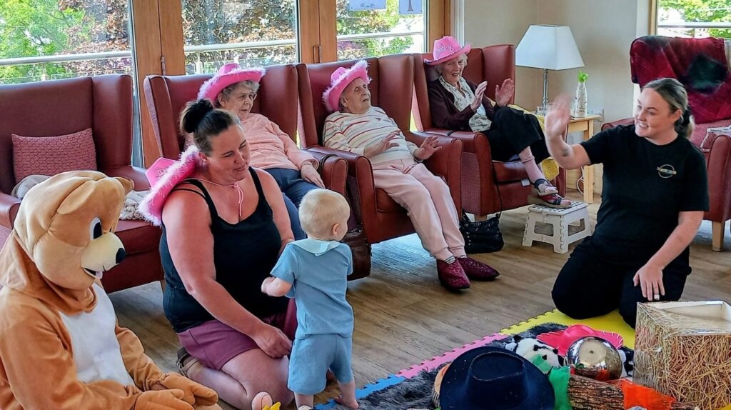A group of elderly women in pink hats sit in armchairs, whilst a woman, a toddler, and someone dressed as a bear play with toys on the floor. Another woman sits close by, waving and smiling.