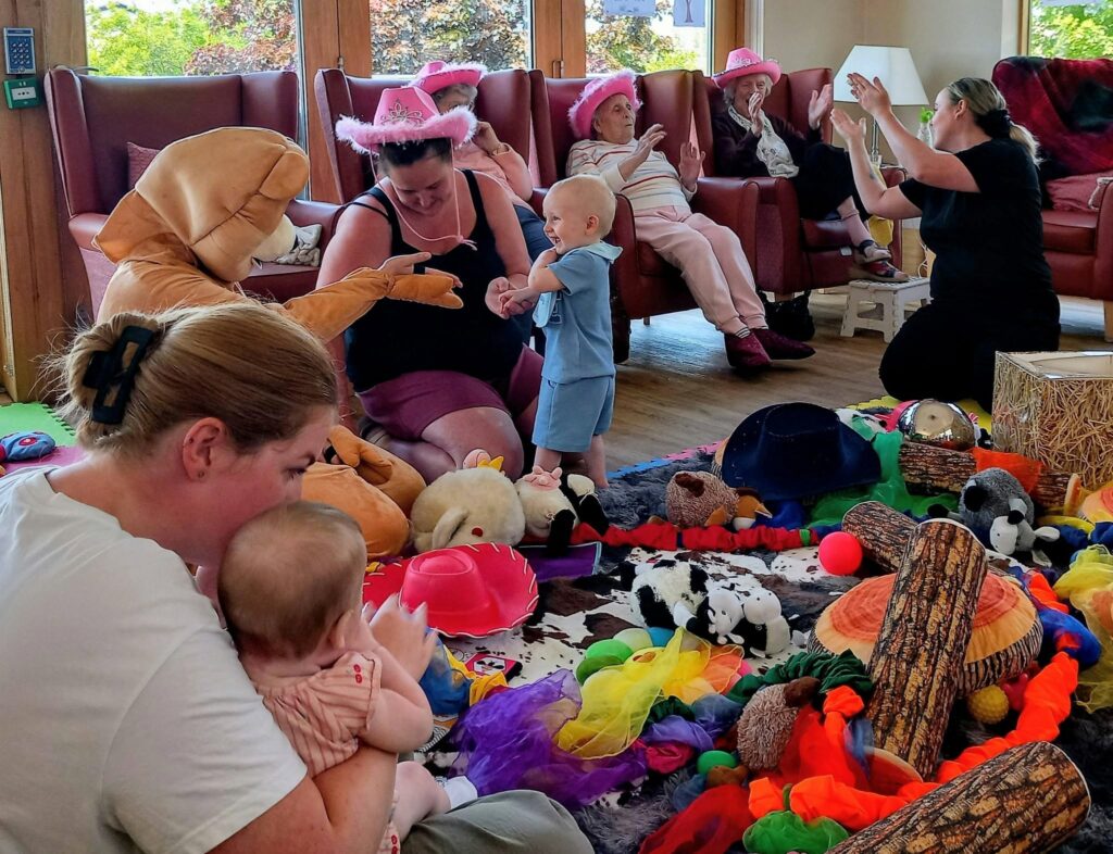 Adults and children sit and play together in a colourful room filled with toys. Some adults wear pink cowboy hats, and one is in fancy dress. A woman leads a group activity as others watch and join in.