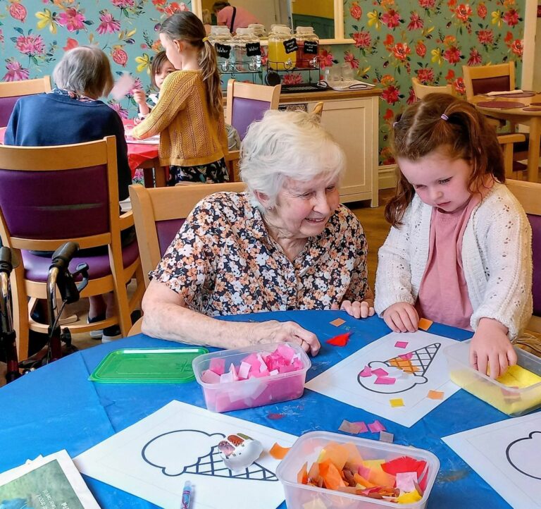 At Lear House’s Craft Morning, an elderly woman and young girl sit together making colourful ice cream-themed crafts—Little Hands & Big Hearts creating memories as others join in the creative fun in the background.