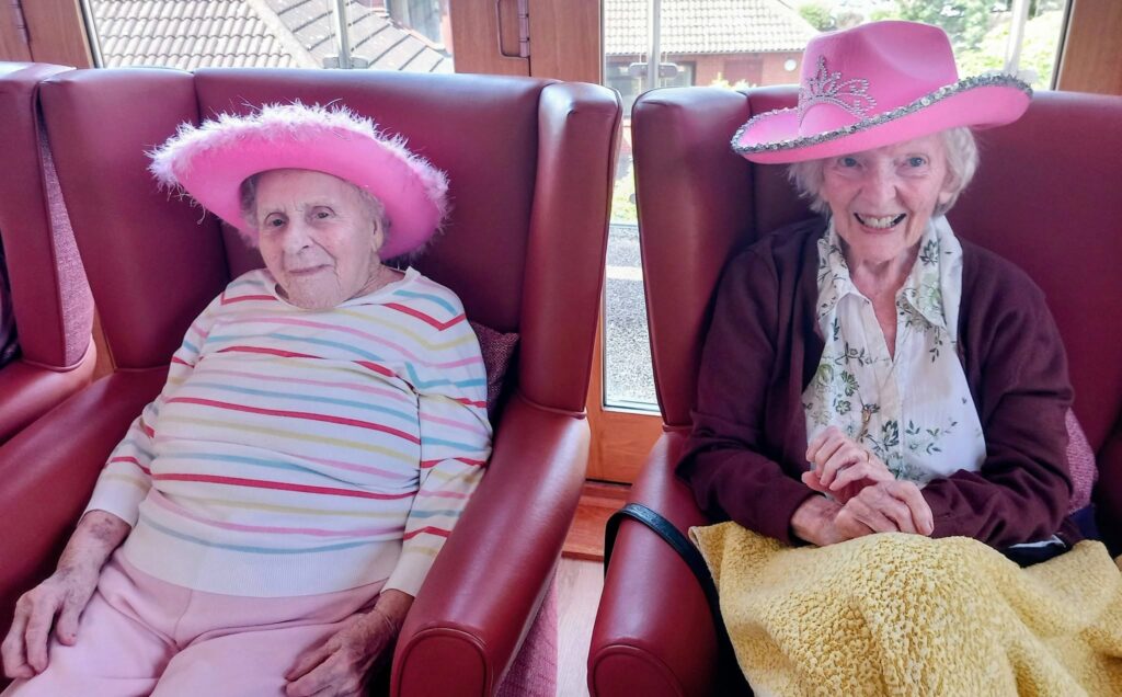 Two elderly women sit indoors in red chairs, both wearing pink hats. The woman on the left wears a striped jumper and light trousers; the one on the right has a brown cardigan, floral blouse, and yellow blanket on her lap.