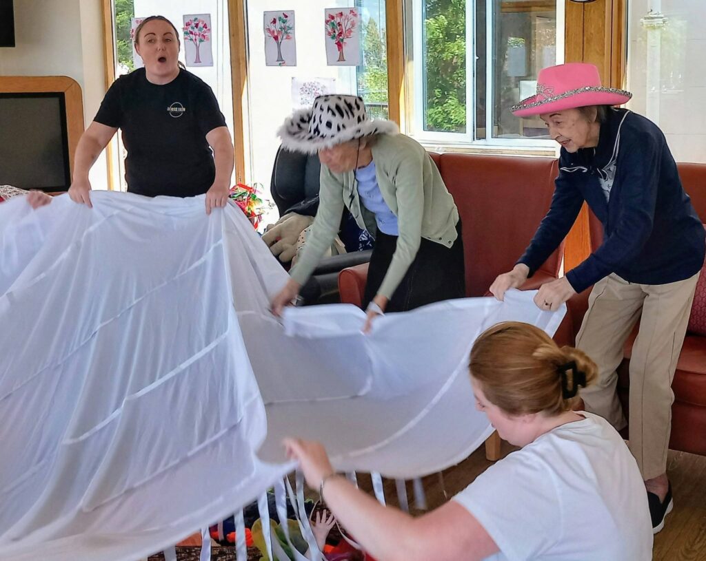 Four women, some in hats, are indoors holding up a large white sheet. Colourful paper art decorates the wall behind them, and natural light streams in through a window.