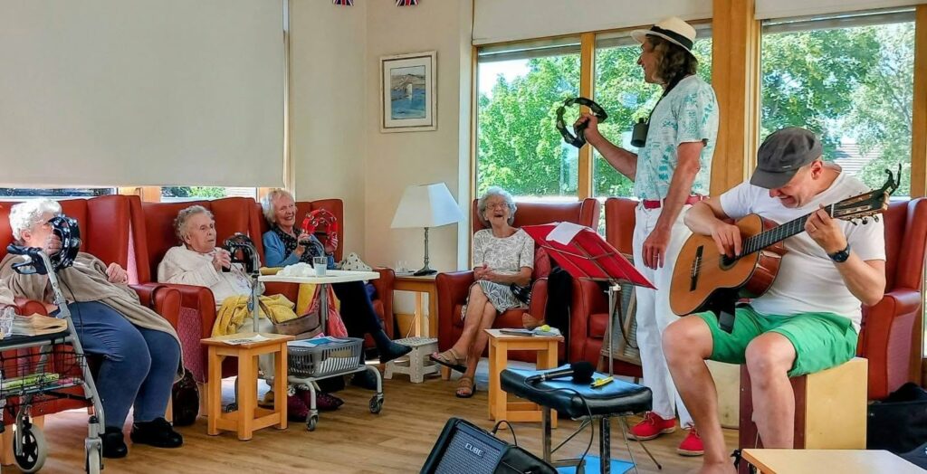 Marko plays guitar as Andrew stands with a tambourine, leading a lively afternoon music session for elderly adults relaxing in armchairs and using walking frames in a bright, homely room.