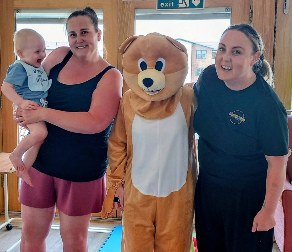 Three adults, including one in a bear costume, stand indoors. A woman is holding a smiling baby. All appear to be at an informal gathering with natural light streaming through windows in the background.