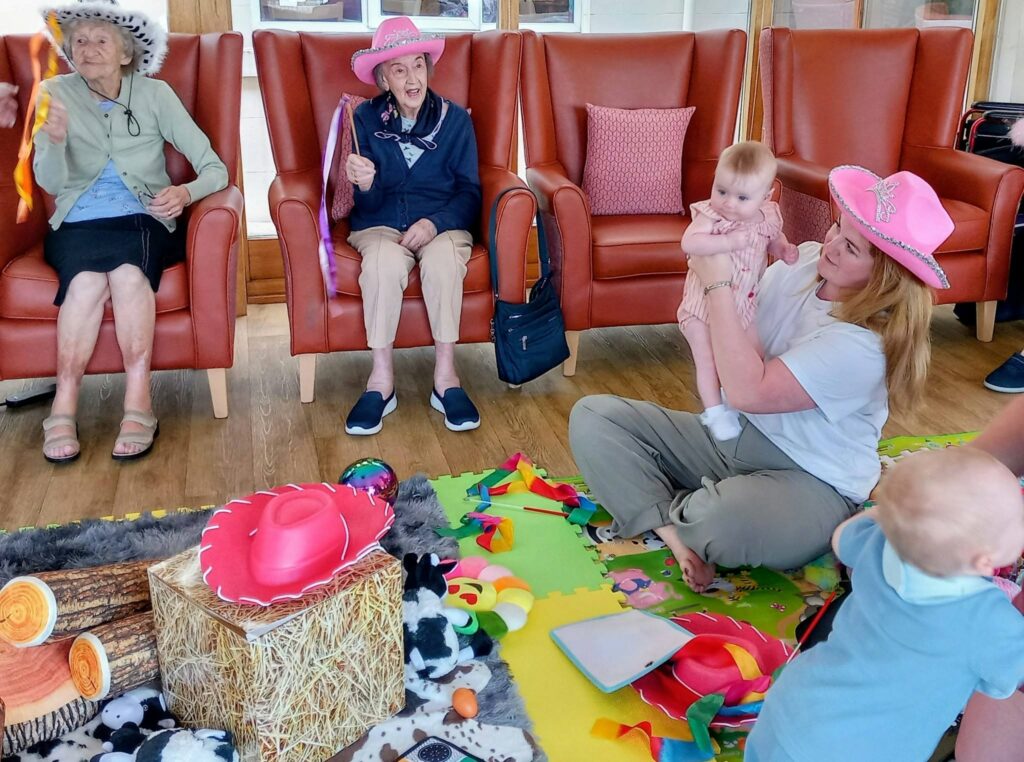 Two elderly women in hats sit in armchairs, while a woman in a pink hat holds a baby on a play mat scattered with toys, beside another baby. The setting looks like a communal room.
