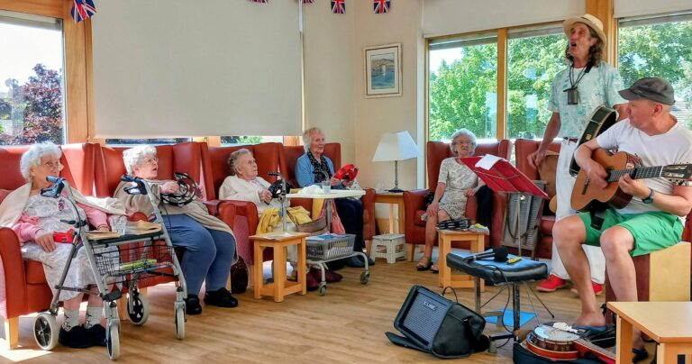 On a sunny afternoon, Marko and Andrew entertain elderly residents in a lounge decorated with British flags, as guitar music and singing fill the room.