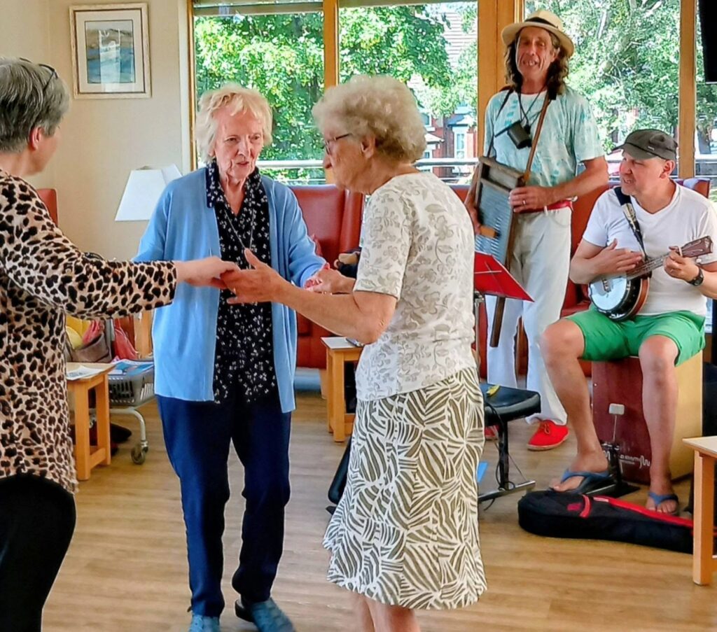 Two elderly women dance together on a splendid afternoon, as Marko strums a banjo and Andrew plays the guitar, filling the sunlit room with music whilst trees sway outside the tall windows.