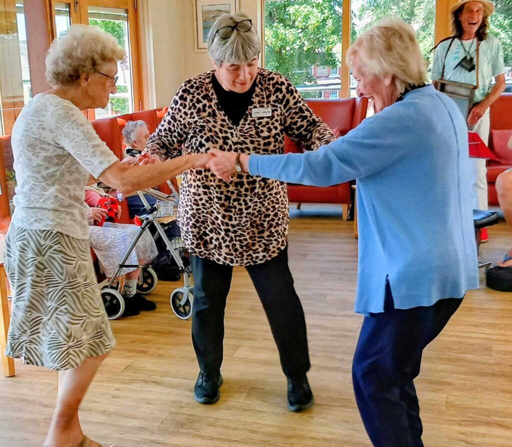 Three elderly women dance together in a brightly lit room with large windows, enjoying a lovely afternoon. Others and a walking frame are visible; the women hold hands and smile warmly.