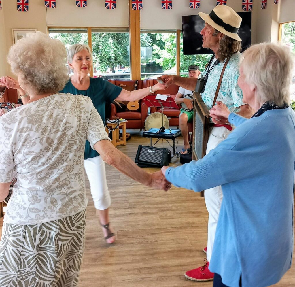Marko and Andrew join two others, holding hands and dancing in a circle on a lively afternoon in a decorated room with British flags and musical equipment in the background.