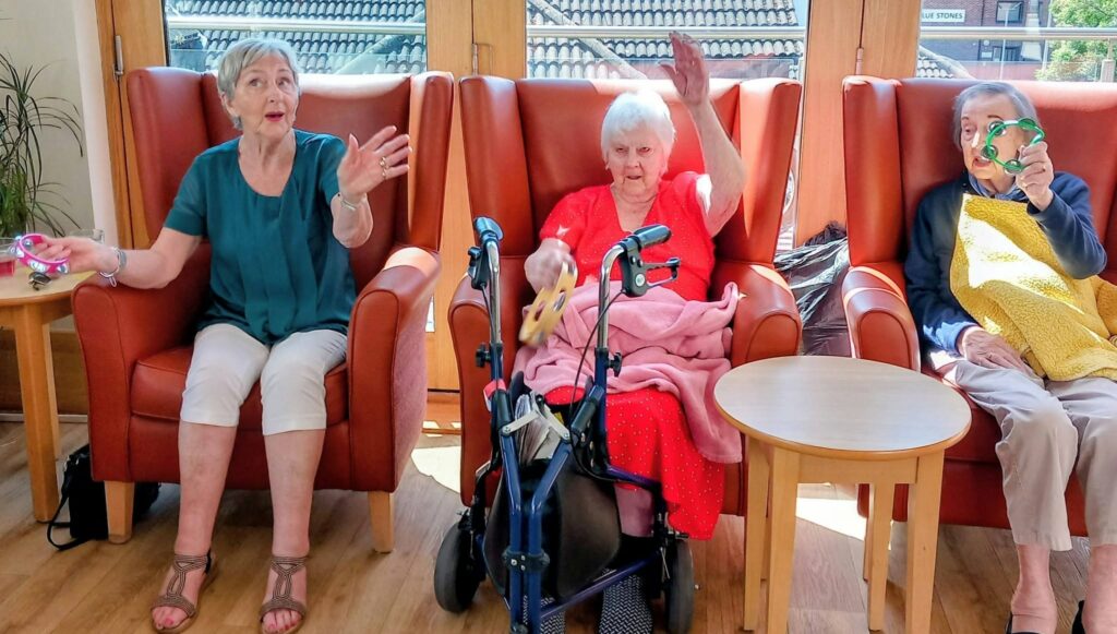 Marko and Andrew, two elderly friends, sit on armchairs indoors on a sunny afternoon, each holding small musical instruments as a third person with a yellow blanket enjoys playing the tambourine.
