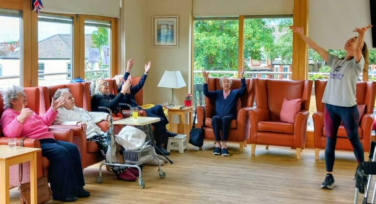 A group of elderly people in armchairs raise their arms whilst a Caroline's Popcats instructor leads an energetic exercise class in a bright, spacious room with large windows.