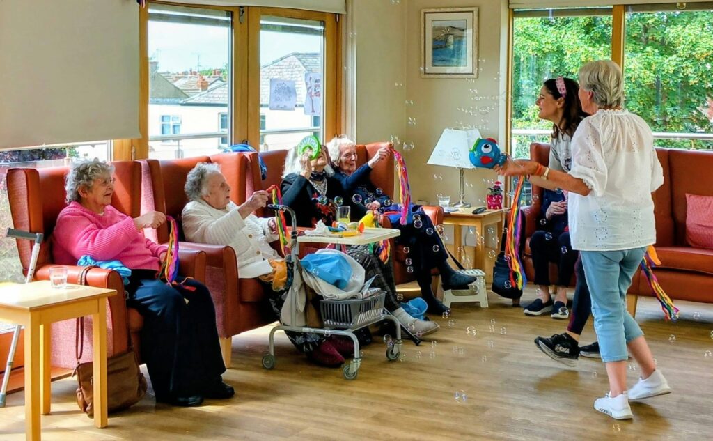A group of elderly women sit on sofas in a bright room, engaging with two younger women leading a ribbon and bubble activity. A walking frame, drinks, craft supplies, and puzzles are visible on a table.