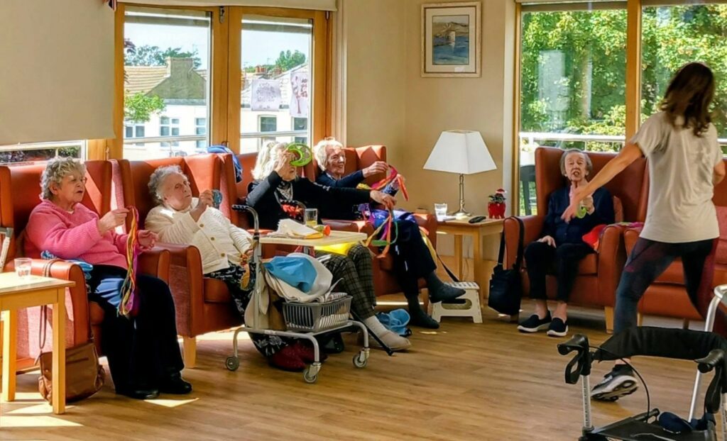 Five elderly women sit in chairs, holding colourful rings and ribbons, enjoying a seated exercise activity that blends gentle dancing and movement, led by a standing instructor in a sunlit room with large windows.