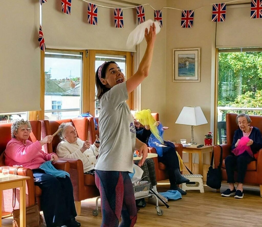 A woman leads a seated dance activity for four elderly adults in a well-lit room adorned with UK flags.