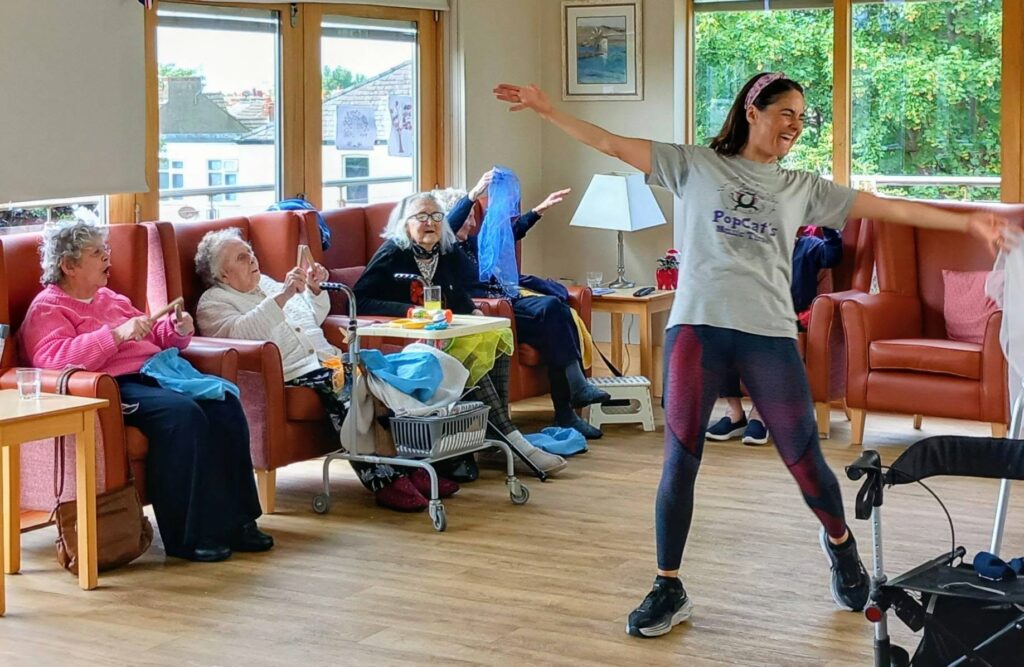 A woman leads a lively seated dance class for elderly people in a bright room with armchairs. Some participants wave their arms, whilst others watch and smile.