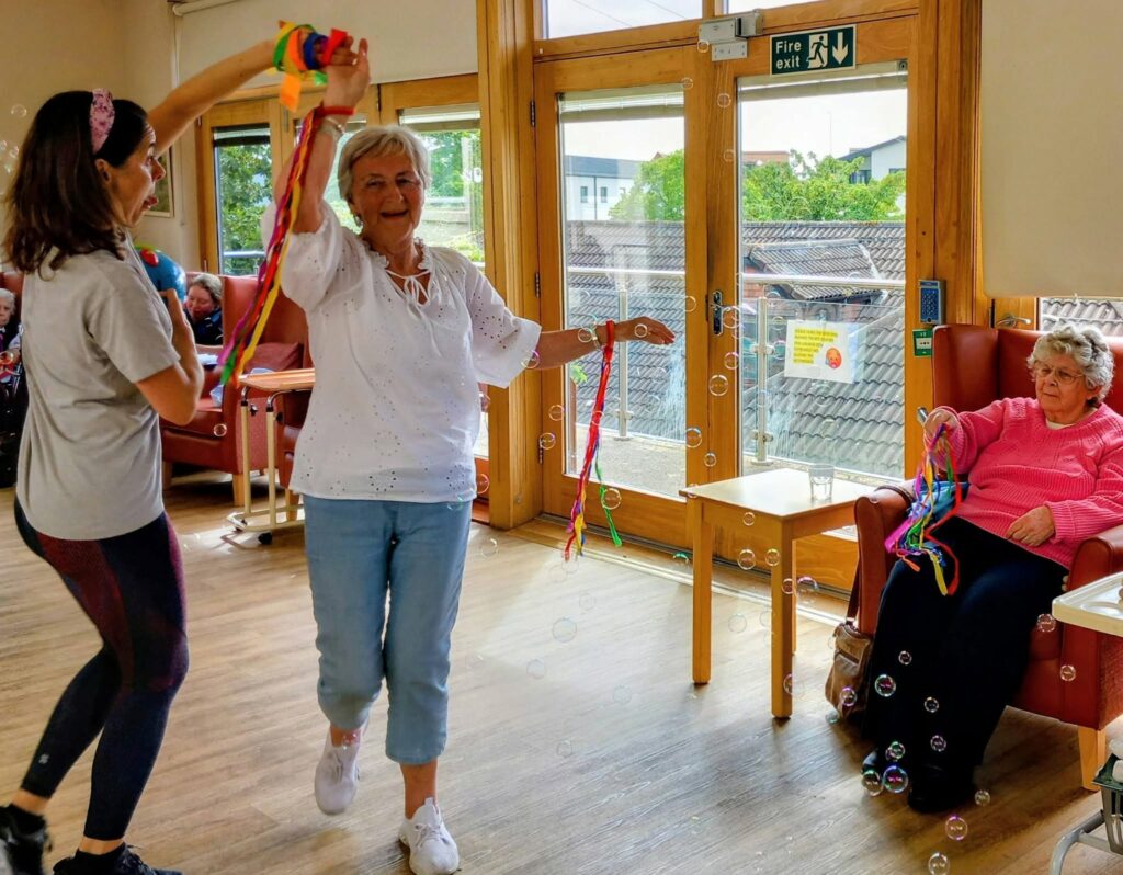 Two women dance with colourful ribbons and bubbles in a bright room, as another woman in pink sits watching. Others by large windows enjoy the lively performance set against buildings and greenery.