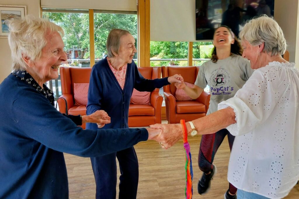 Four women stand indoors in a circle, holding hands and smiling as though dancing. One wears a “PopCat’s Music Time” top. Orange chairs and large windows add to the cheerful atmosphere.