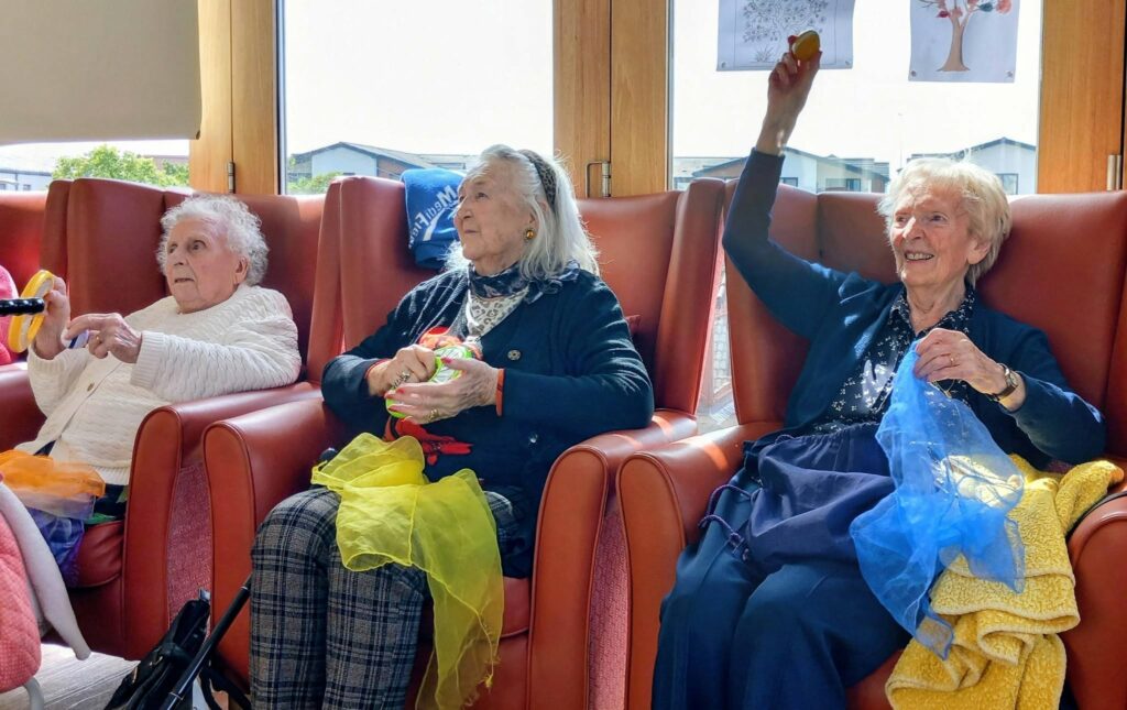 Three elderly women sit in armchairs, smiling and holding colourful scarves and items, taking part in a percussion activity in a bright room with large windows.