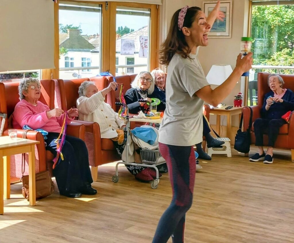 A woman energetically leads a dance activity in a bright room as elderly people seated on orange chairs watch and join in, some holding colourful ribbons.