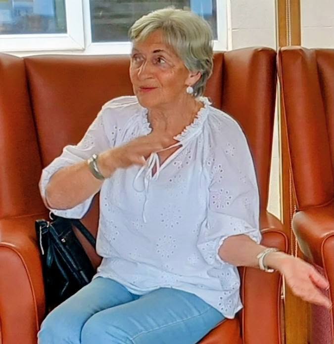 An older woman with short grey hair sits on a brown chair, dressed in a white blouse and light blue jeans. She gestures as if explaining puzzles, with a black bag beside her.