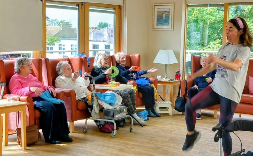 A woman leads an exercise session in a common room, guiding several elderly women who watch, join in, and dance along from armchairs—some holding small hand weights.