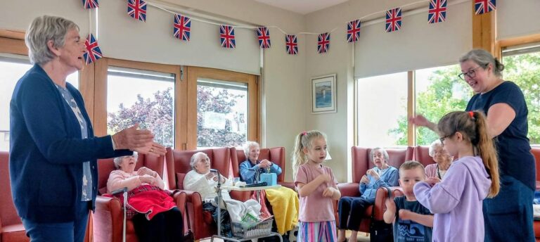 Children and adults share stories and sing songs whilst the youngest visitors play nearby, as elderly guests watch from armchairs by large windows beneath Union Flag bunting.