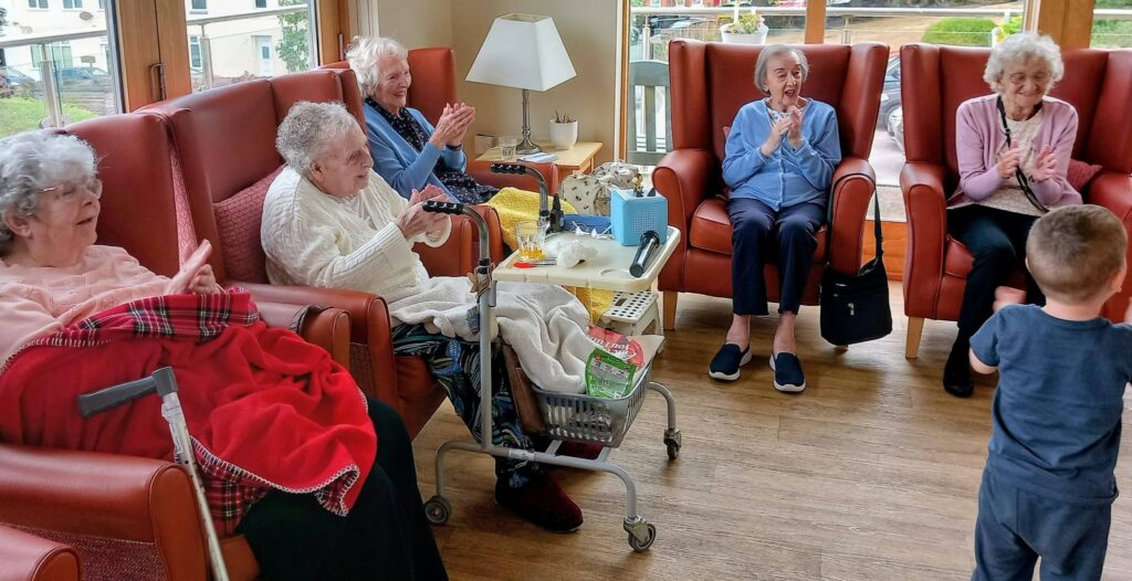 A group of elderly women sit in armchairs, clapping and smiling as one of their youngest visitors shares stories and songs in a brightly lit room with large windows.